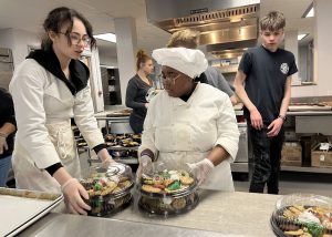 Two culinary students dressed in white kitchen garb prepare to package plates of cookies for sale.