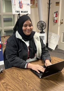 A young woman wearing a dark headscarf and dark winter coat sits and types on a laptop computer.
