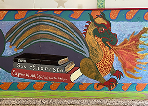A brightly colored photo of a mural painted on a school wall featuring a dragon that is breathing fire and sitting on a pile of books. 