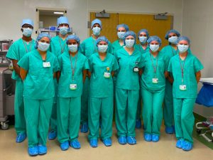 Several individuals stands in two rows, wearing surgical garb that covers their clothing, face and hair.