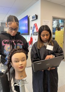 Two students, one with a laptop and another working on the hair of a mannequin, prepare for a class exercise in the cosmetology program. 