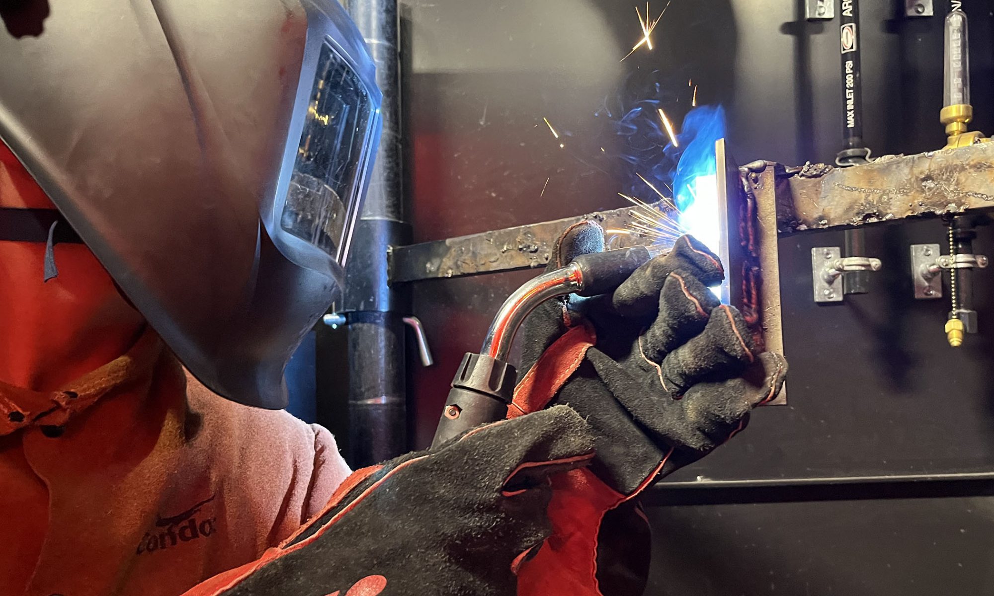 A student covered by a welder's mask and safety gear ignites uses his torch to piece together his metalwork.