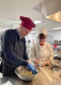 A man in a dark apron and red chef's hat works at hand mixing ingredients in a bowl as a student in traditional chef's garb watches on.