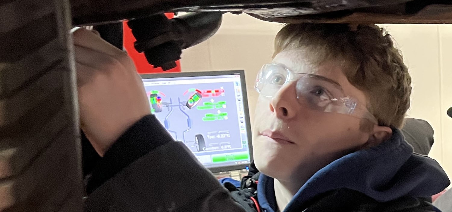 A student in safety goggles works on the undercarriage of a car that is lofted up inside a garage.