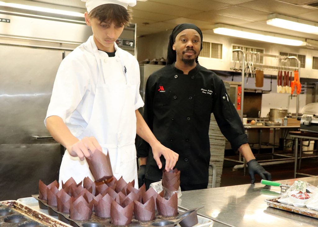 A chef and his student prepare a dish from inside the kitchen.