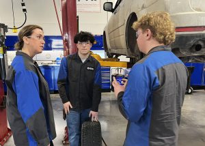 Two students and a school principal share a conversation inside the garage next to a car hoisted on a jack.