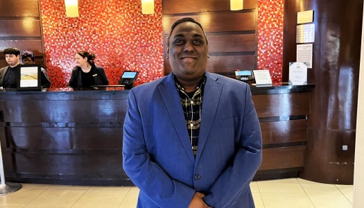 Young Black man in a blue suit stands in front of a customer service desk at his hotel.