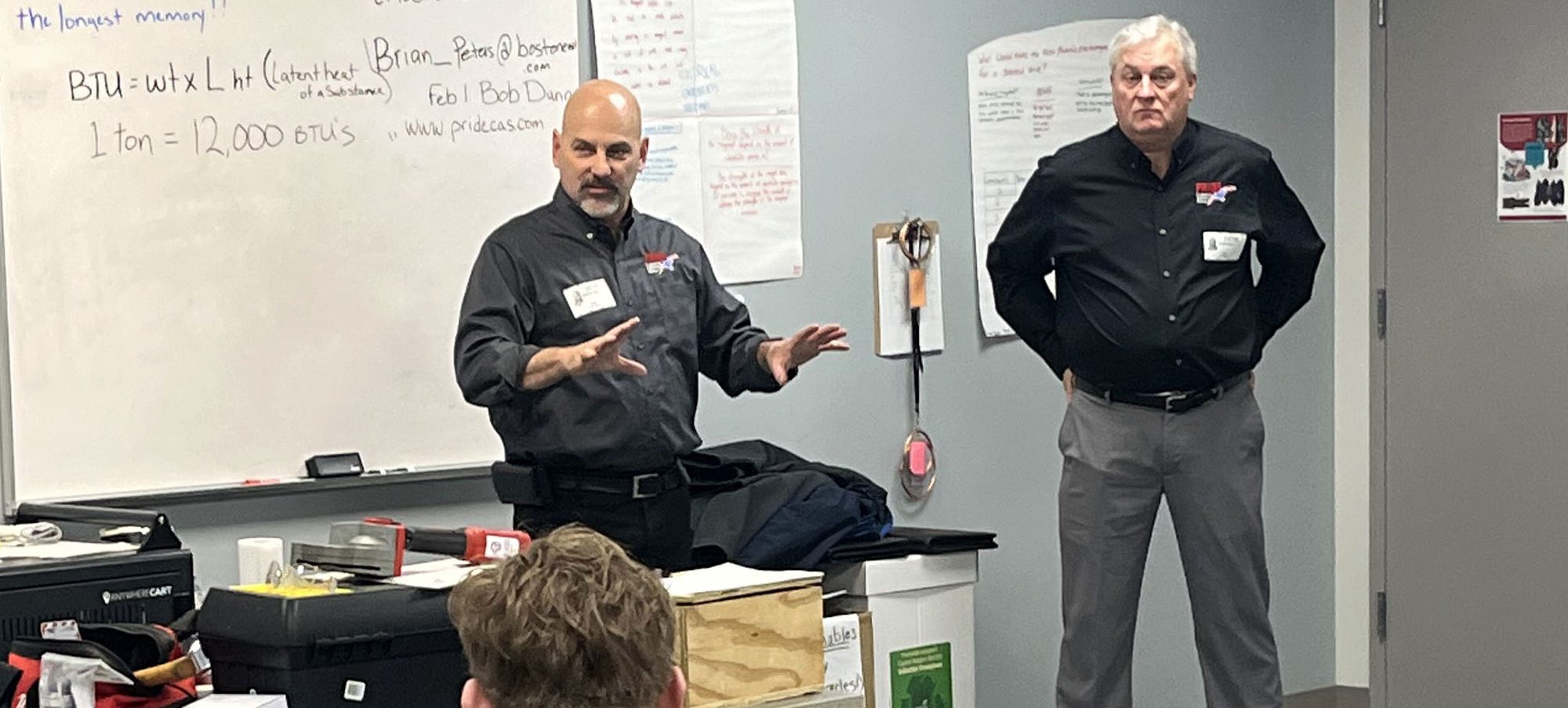 Two gentlemen in dark company shirts speak before a classroom of students.