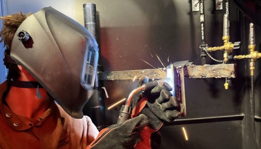 A student covered by a welder's mask and safety gear ignites uses his torch to piece together his metalwork.