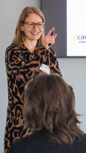 A female presenter smiles as she points to a student off camera. Another student sits in the foreground with the back of his head towards the camera.