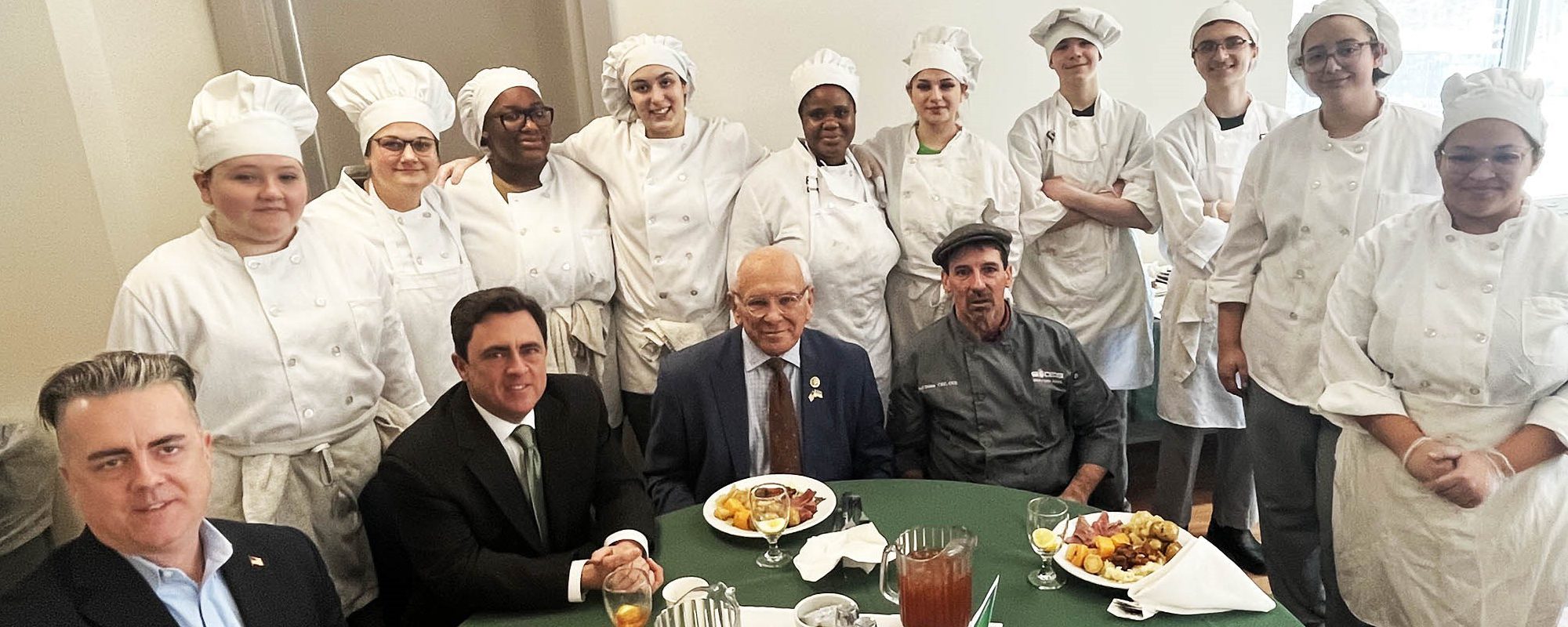 A dozen culinary students in white aprons and hats stand behind politicians who were served food on top of a table draped with a green tablecloth.