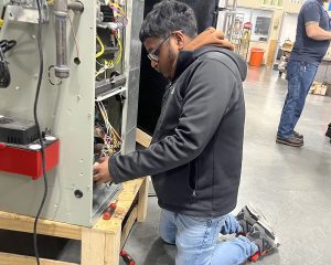 A student in a dark jacket works on a piece of equipment, exposing wires and infrastructure for an HVAC system.