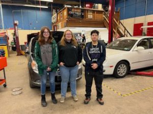 Three young women stand to pose together for a picture inside the school's automotive repair garage.