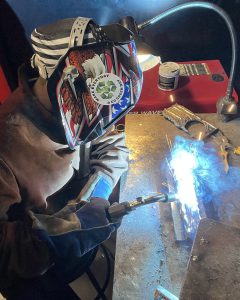 A student in a welders helmet is in the midst of welding metal, her torch lit and emitting a bright white and blue glow.