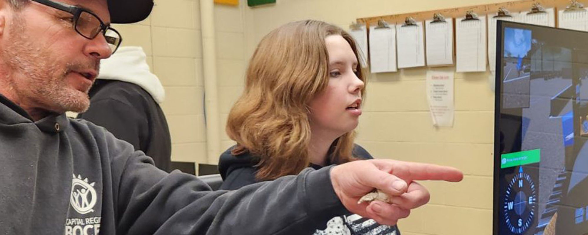 A male teacher in a baseball cap is seated next to a young female student as he points to a computer monitor.