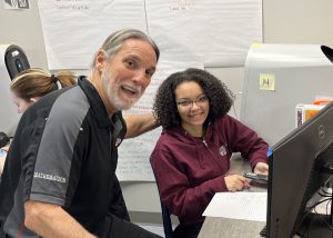 A male teacher rests his hand on the chair behind the back of a female student's head as they both turn and smile to the camera.
