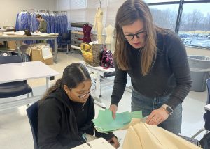 A female teacher stands over the desk of a student as they each look over material in the teacher's hand.