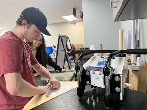 A student in a red shirt and blue baseball cap prepares to print a T-shirt as another student looks on.