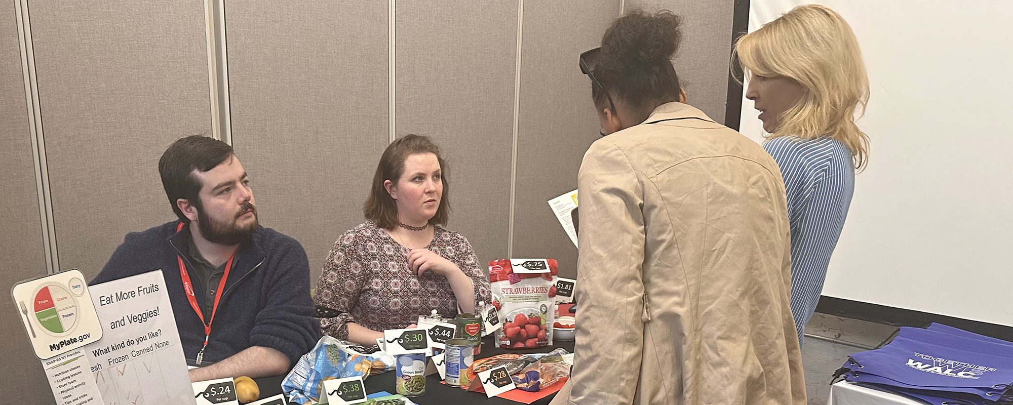 Two people seated behind a display table talk to two others.