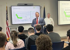 Students sit in attendance listening to a man in a suit, a politician, as he speaks in front of a chart projected behind him.