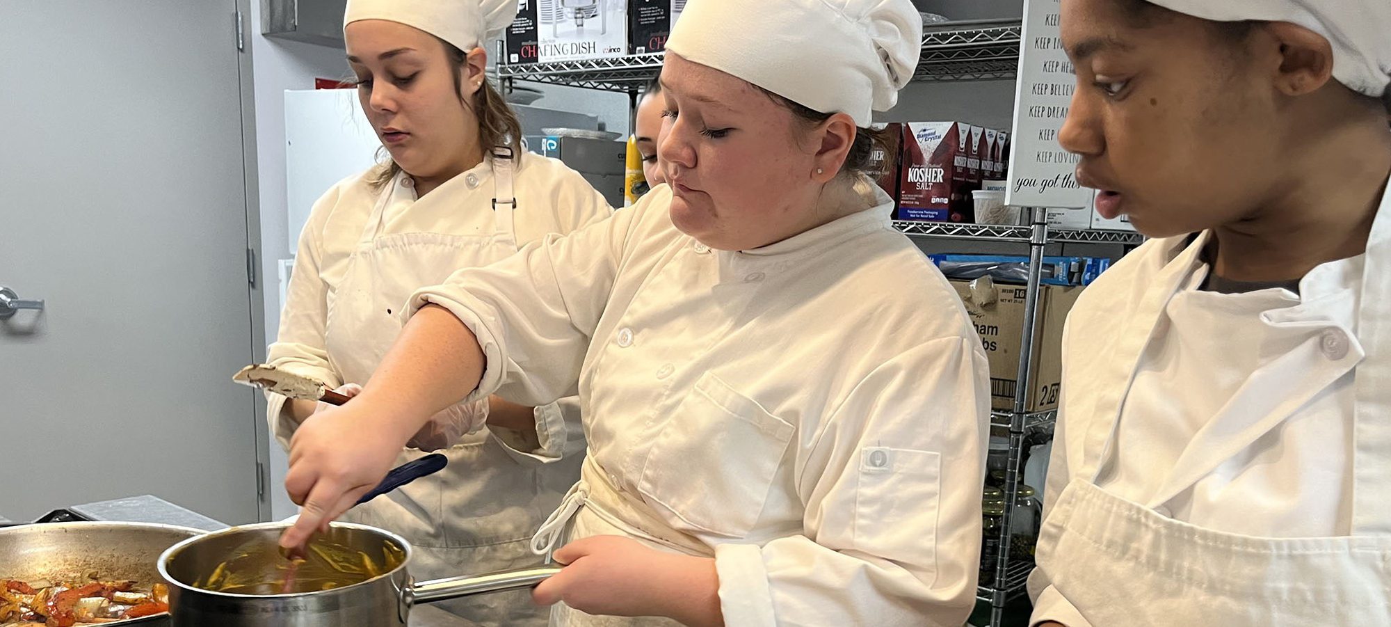 A culinary student stirs a pot as three more, all dressed in white, look on.