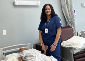 A student dressed in blue scrubs smiles for the camera while tending to a mannequin patient.