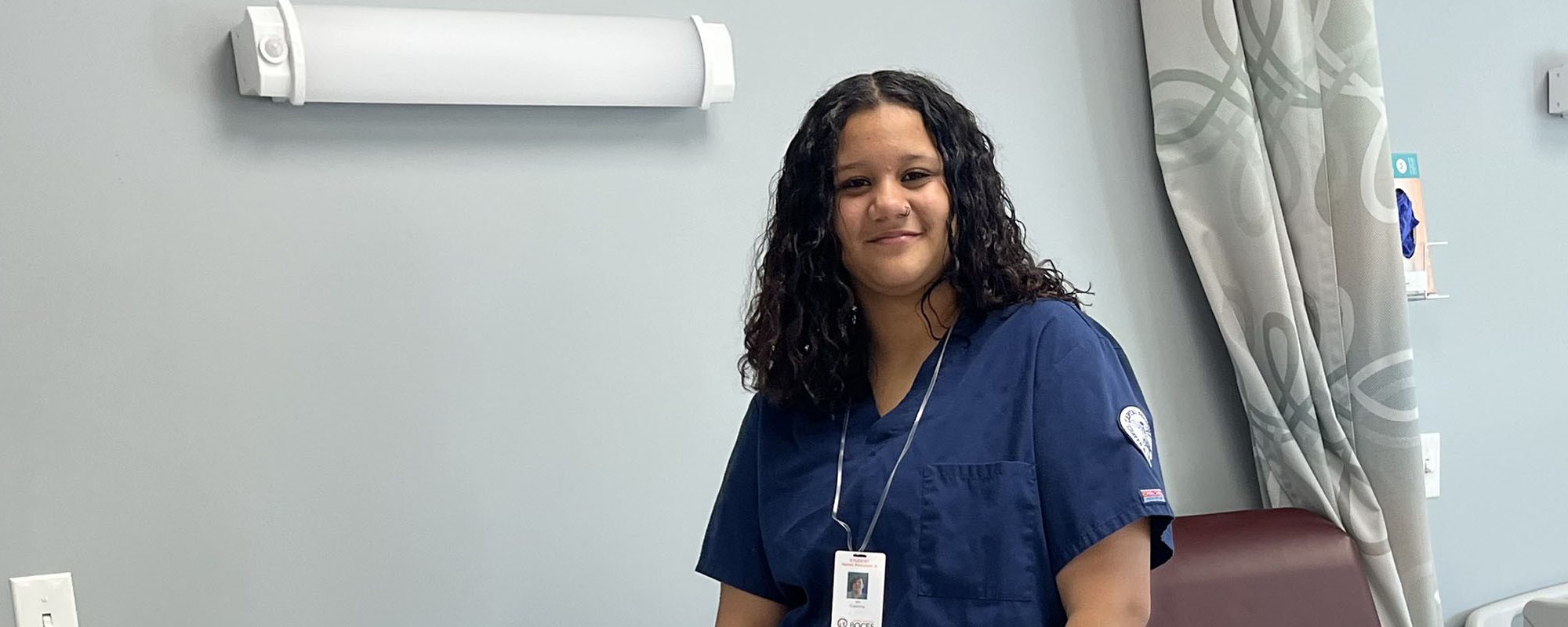 A student dressed in blue scrubs smiles for the camera while tending to a mannequin patient.