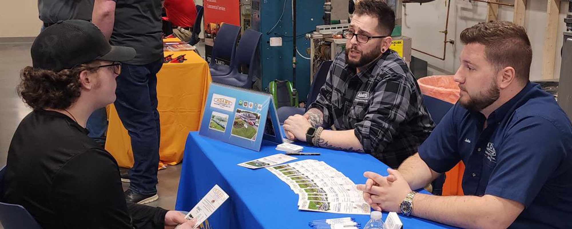 A student sits in front of a table, listening to two representatives from a prospective employer at a job fair.