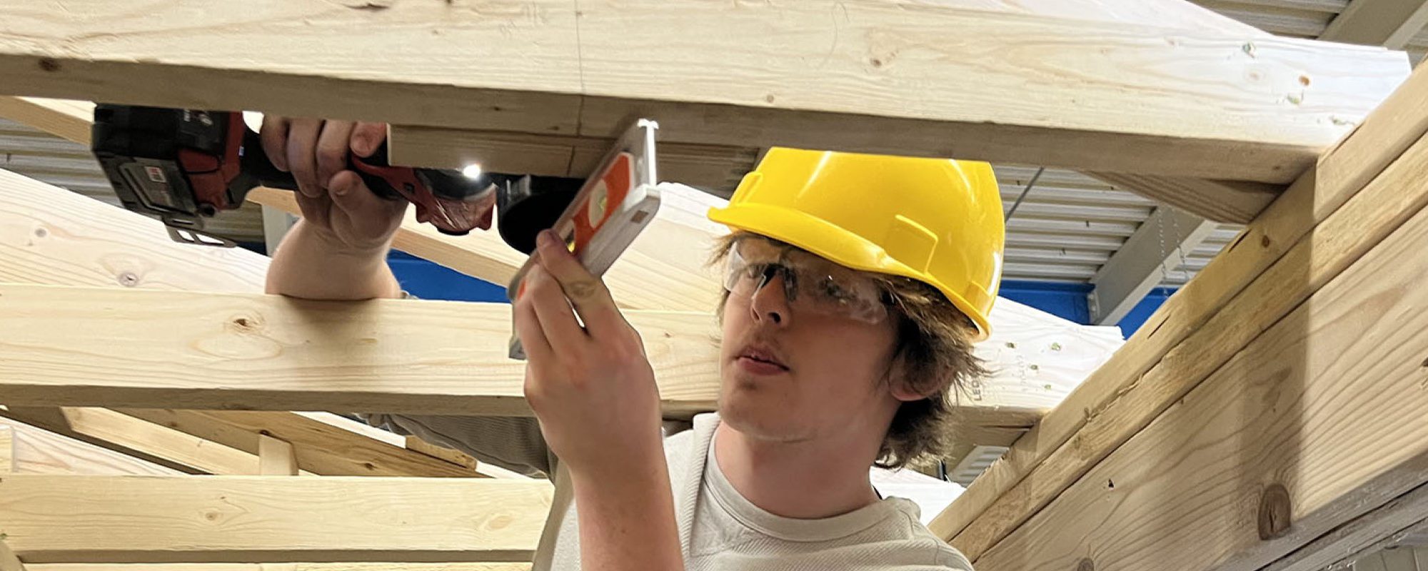 A student in a yellow hard hat holds a level before fixating an assembly to a wooden frame.