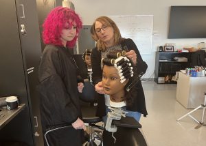A student takes instruction from a teacher on how to apply hair rollers on a mannequin's head.