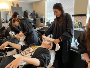 A student in a black robe pampers another laying back in a chair with a facial treatment and warm towel.