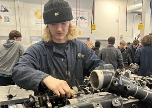 A student in blue coveralls and a cap leans over an engine block.