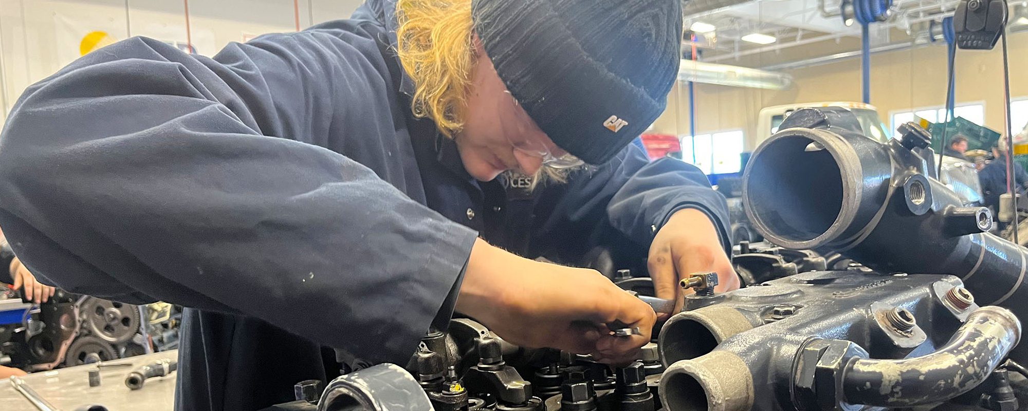 A student in blue coveralls and a cap leans over an engine block.