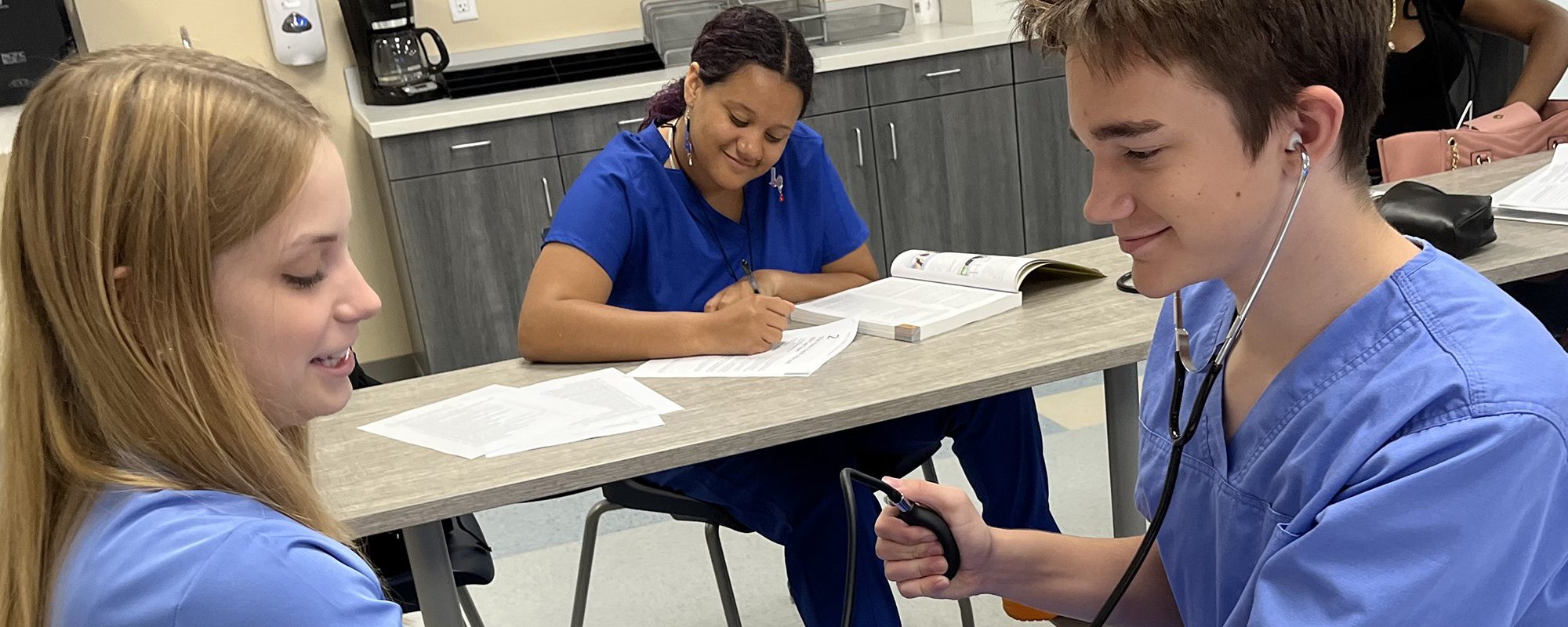 One student listens to another's heartrate while checking that person's blood pressure during a classroom exercise. Another student smiles while taking notes in the background.