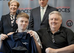 A student holds up a shirt that brandishes the name of his future employer during a Signing Day ceremony. He is surrounded by three members of the school community.