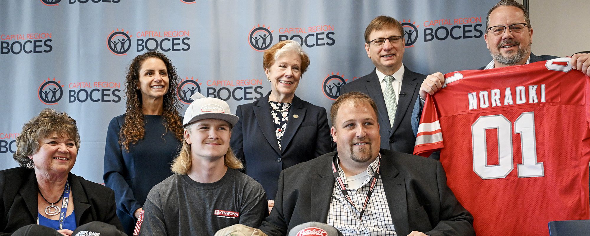 A student wears a baseball cap brandishing the name of his future employer as he's surrounded by school staff and his future boss during a Signing Day ceremony.