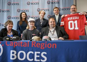 A student sits behind a table wearing a baseball cap that brandishes the name of his future employer. He is surrounded by members of the school community.