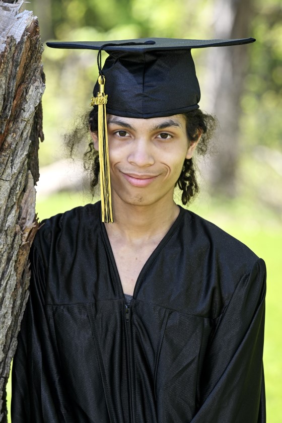 a student stands with a tree dressed in cap and gown