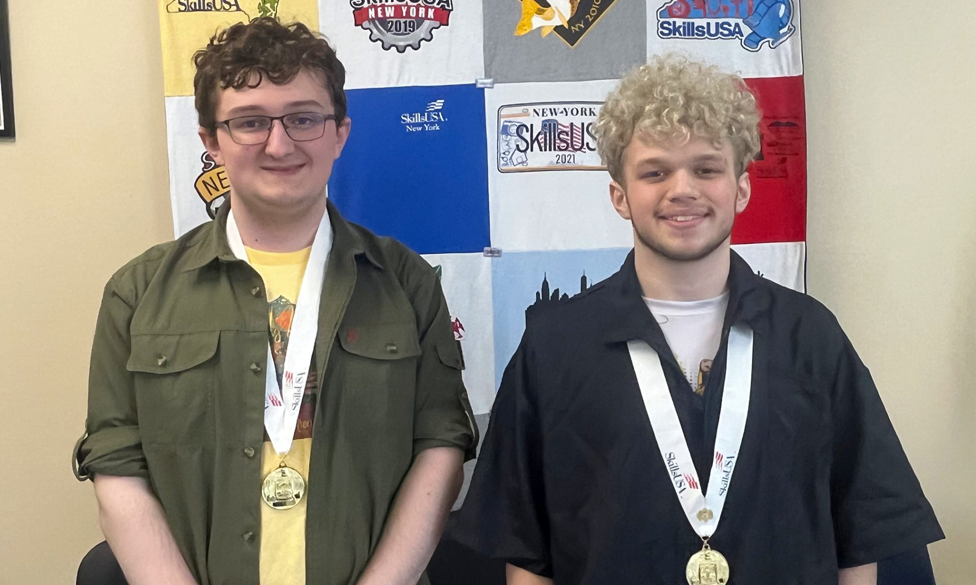 Two students are smiling at the camera, standing next to each other and each one is wearing a medal around the neck.