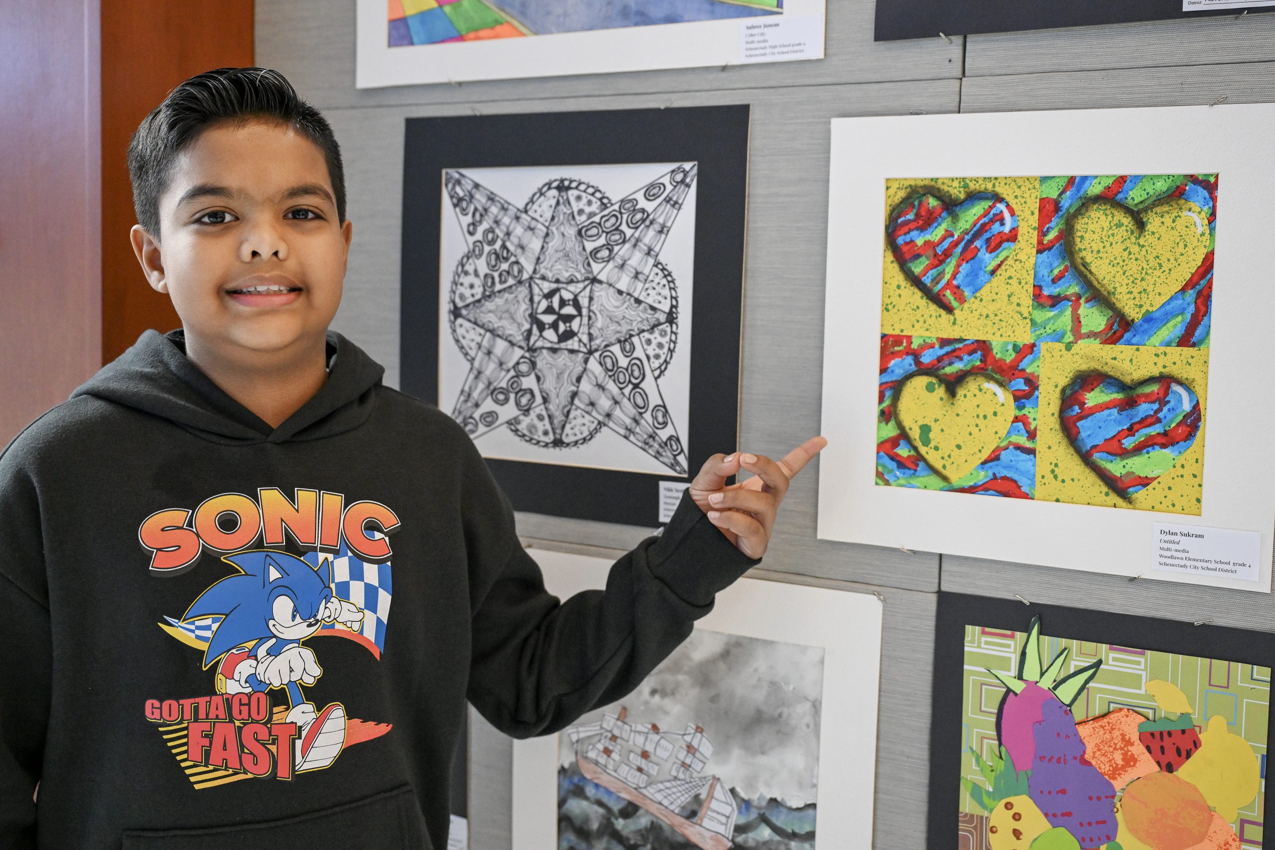 A student is standing next to a wall where students' artwork is hanging. The student is pointing to a piece of art that is colorful and features four hearts