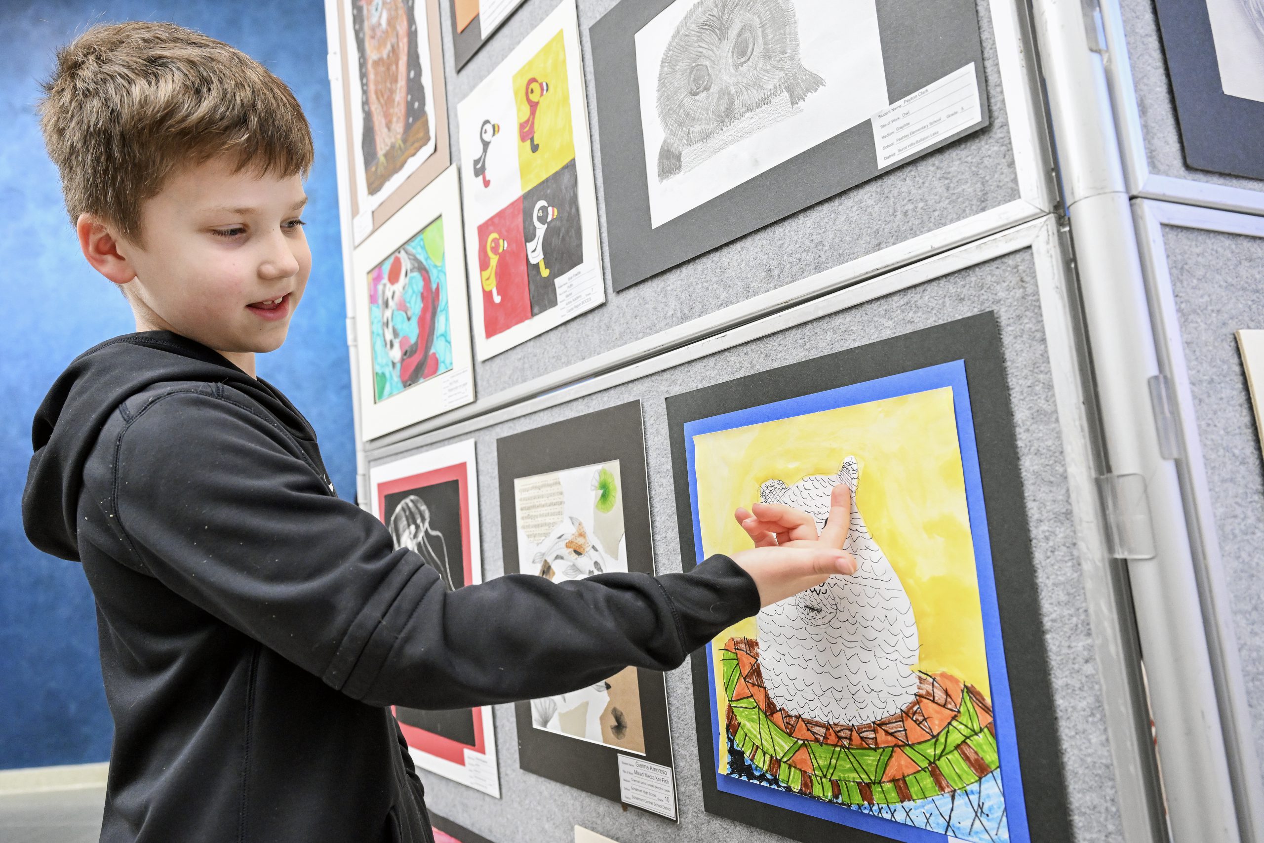 A student is standing next to a wall where students' artwork is hanging. The student is pointing to a piece of art that is colorful