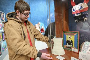 A student stands next to a display of three-dimensional artwork, including a red, white and blue mask hanging on the wall, a while clay piece and others