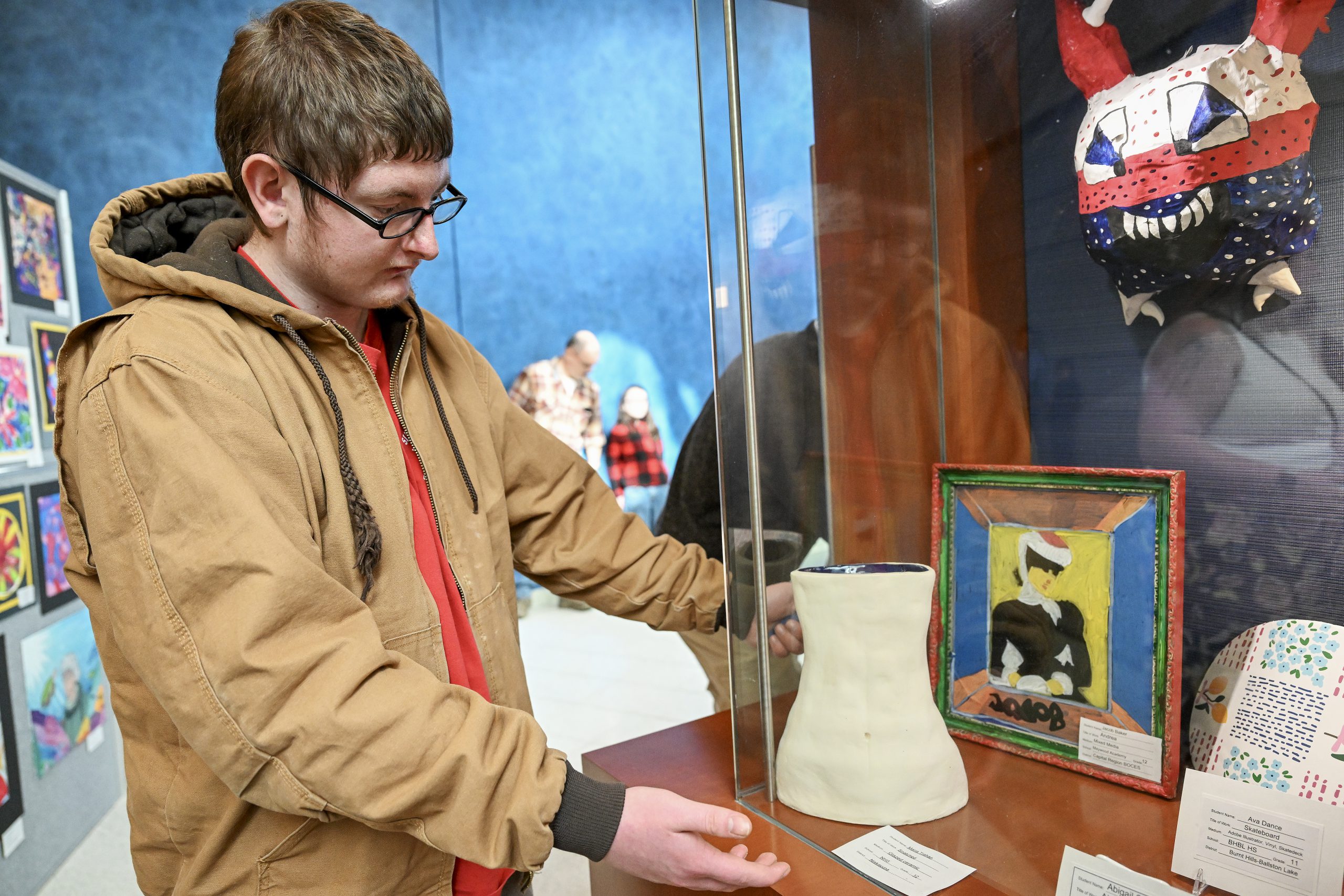 A student stands next to a display of three-dimensional artwork, including a red, white and blue mask hanging on the wall, a while clay piece and others
