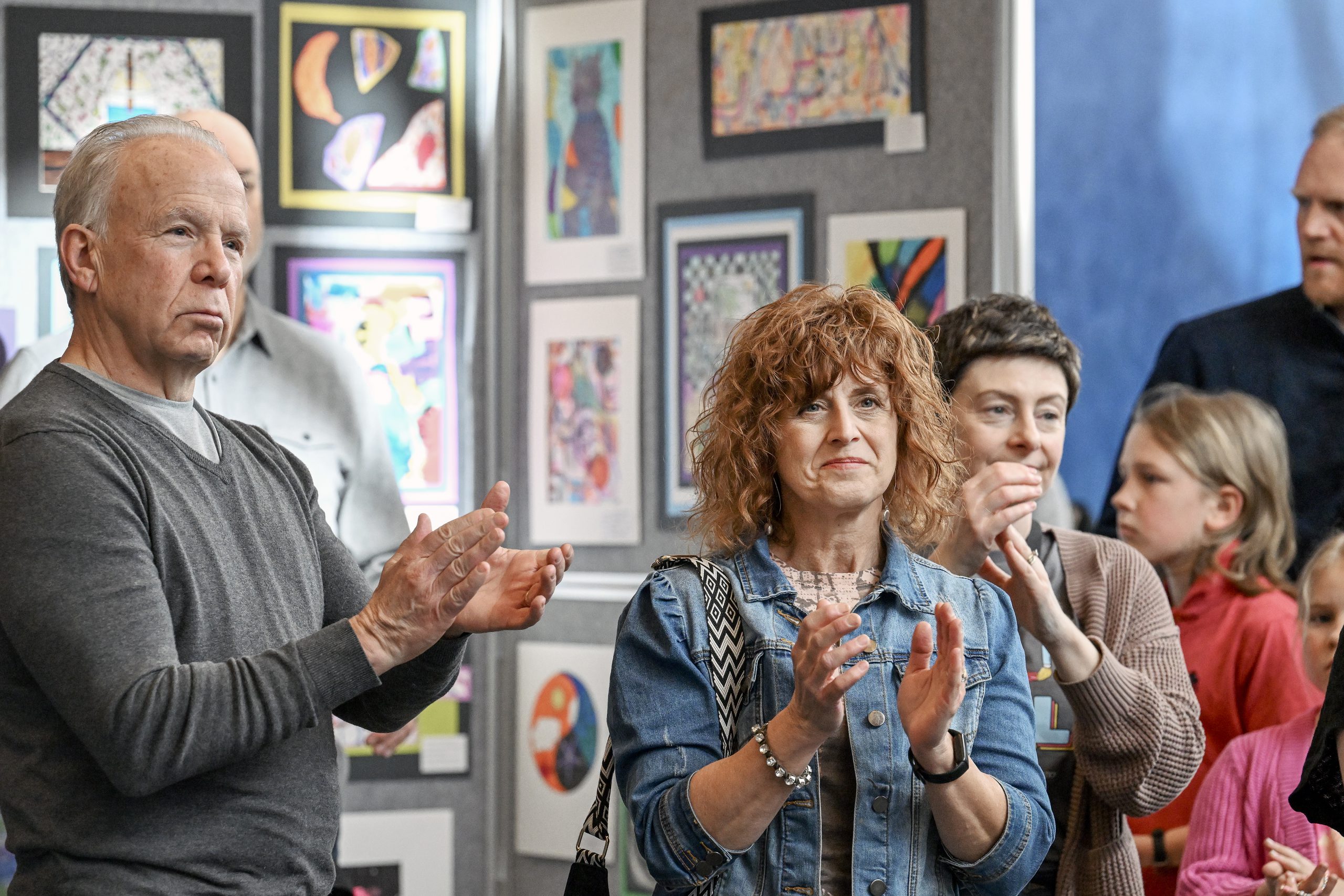 A group of people are at an art show; student art work is hanging on the walls behind the people. At the forefront are two people who are applauding