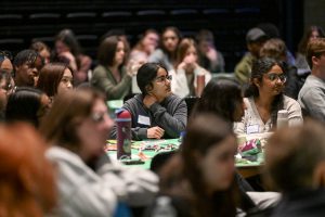 Students sit at round tables listening to a presented (not pictured).