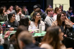 A room full of students sit at round tables listening to a presented (not pictured).