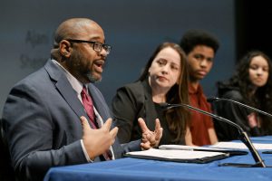 An adult speaks at a presenters table while another adult and two students look on.