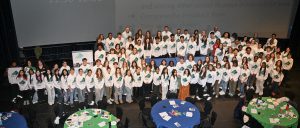 A large group of students wearing white t-shirts stand in a group in an auditorium