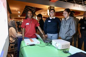 Three students gather at a table with a laptop that is projecting onto a large screen.