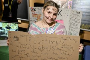 A student holds a sign that indicates "positive labels" while also displaying the word "kind" across her forehead.